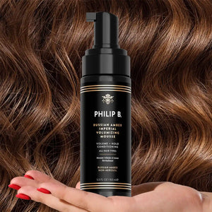 Philip B - Russian Amber Imperial Volumizing Mousse Non-Aerosol in schwarzer Flasche vor welligem braunem Haar mit rotem Nagellack