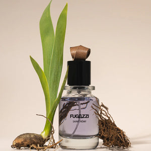 Fugazzi - Saint Rémy Parfumflakon mit violettem Duft, grünem Blatt und Wurzel vor hellem Hintergrund