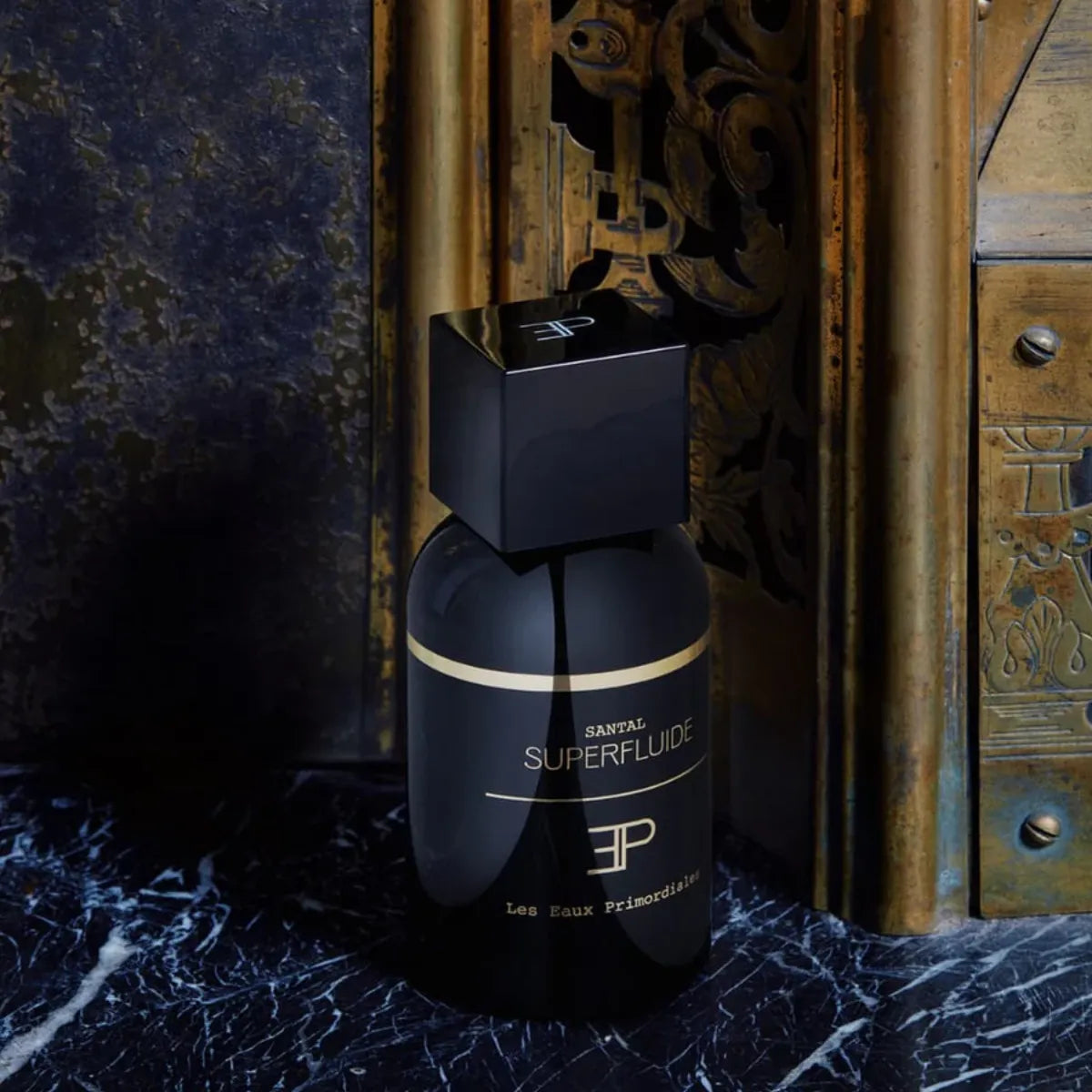 Les Eaux Primordiales - Santal Superfluide schwarzer Flakon mit goldener Linie vor kunstvollem Hintergrund aus dunklem Marmor und Holz