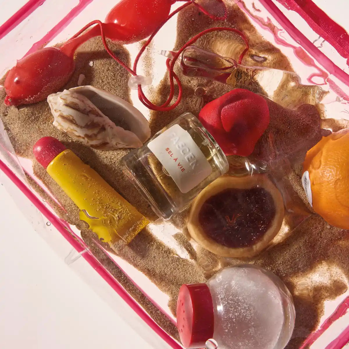 Fascent - Sel à Vie Parfumflakon umgeben von Sand, Muschel, roten und orangefarbenen Objekten in klarem Behälter