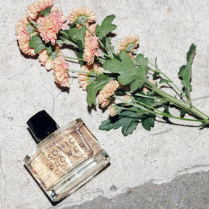 Room 1015 - Sonic Flower Parfumflakon mit zartrosa Blumen auf rauem Steinuntergrund in natürlichem Licht