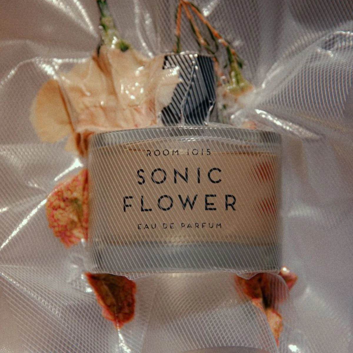 Room 1015 - Sonic Flower Parfumflakon mit floralem Arrangement und transparentem Netz vor unscharfem Hintergrund