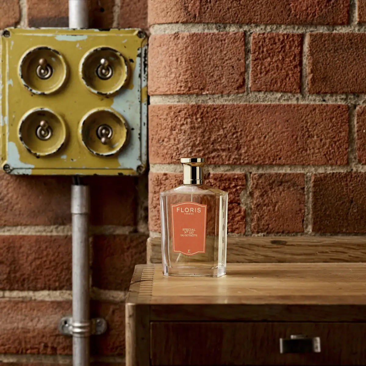 Floris - Special No. 127 Flakon mit goldfarbenem Verschluss und orangefarbenem Etikett auf Holzregal vor Backsteinwand