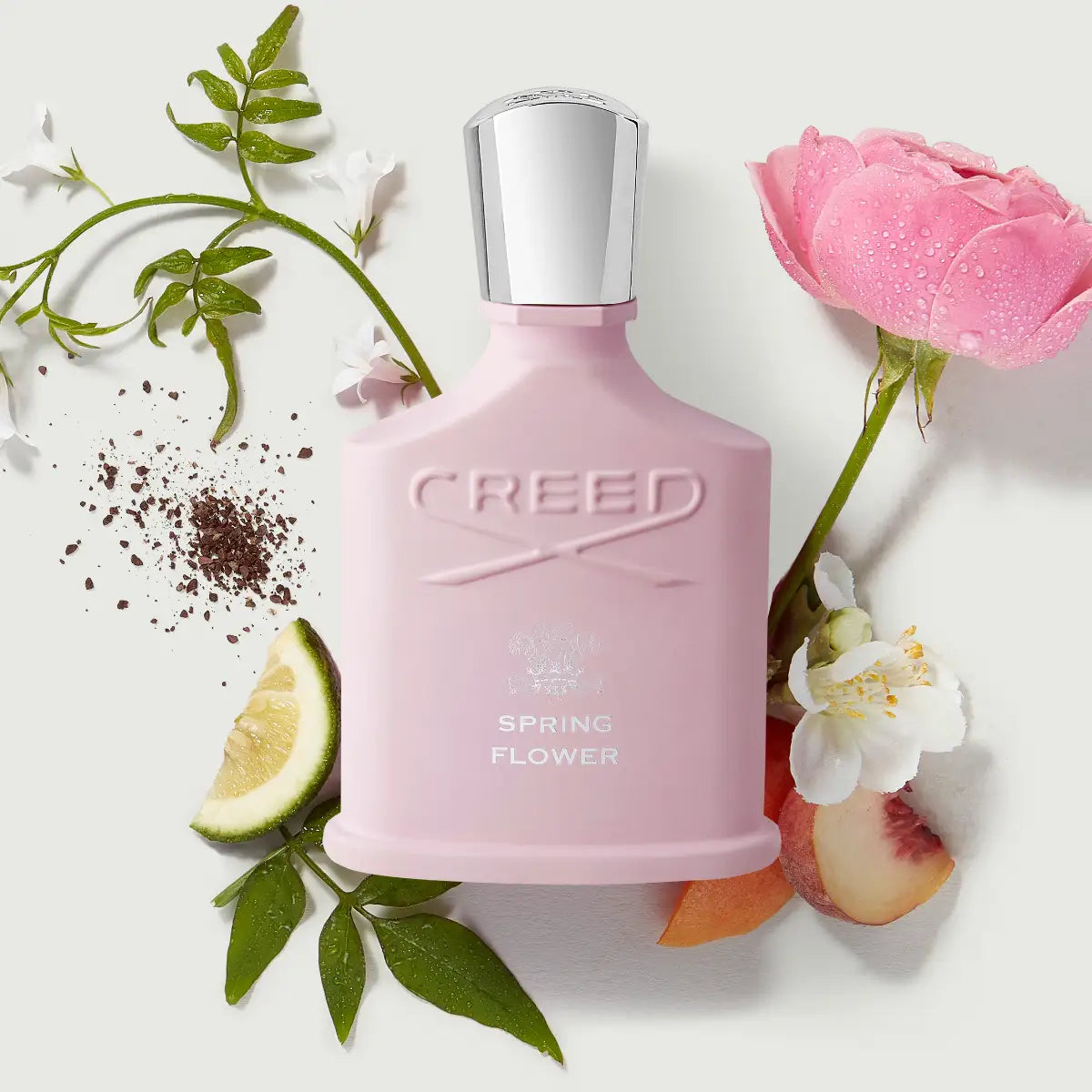 Creed - Spring Flower mattrosa Flakon mit silbernem Deckel umgeben von Blüten und Zitronenscheibe auf weißem Hintergrund