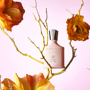 Creed - Spring Flower zartrosafarbener Flakon mit silbernem Verschluss umgeben von getrockneten Blumen auf pastellfarbenem Hintergrund