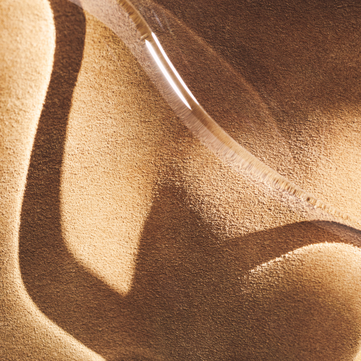 Byredo - Suede - Hand Wash mit transparentem Design vor dezent geschwungenem, sandfarbenem Hintergrund