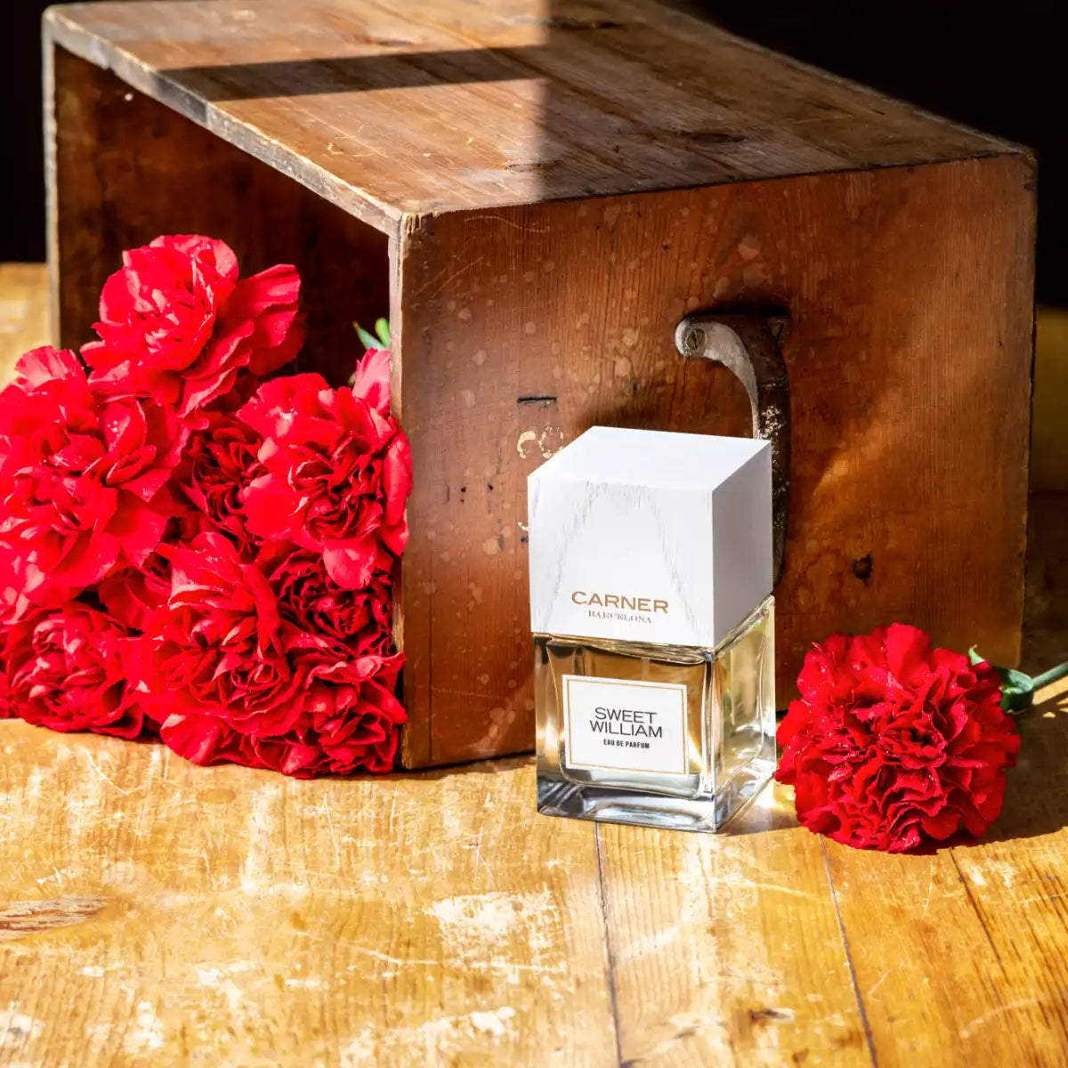 Carner Barcelona - Love Collection - Sweet William Parfumflakon vor Holzbox mit roten Nelken auf Holztisch in warmem Licht