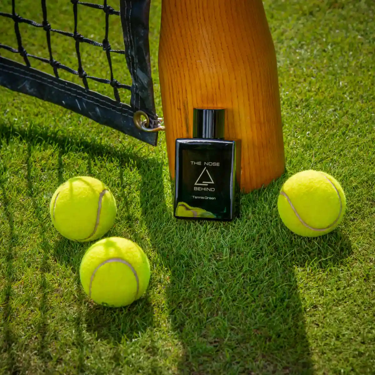 The Nose Behind - Tennis Green Parfumflakon auf grünem Rasen neben Tennisbällen und Holzpfosten im Sonnenlicht