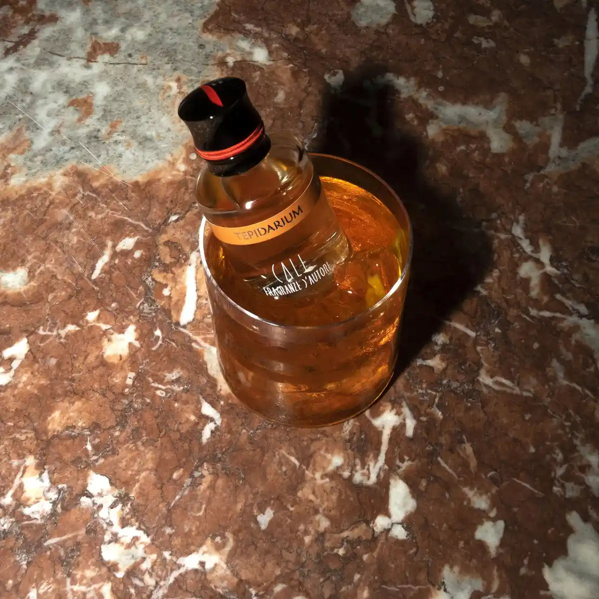 Calé Fragranze d'Autore - Tepidarium Flakon mit orangem Etikett und rotem Band in Getränk auf Steinplatte