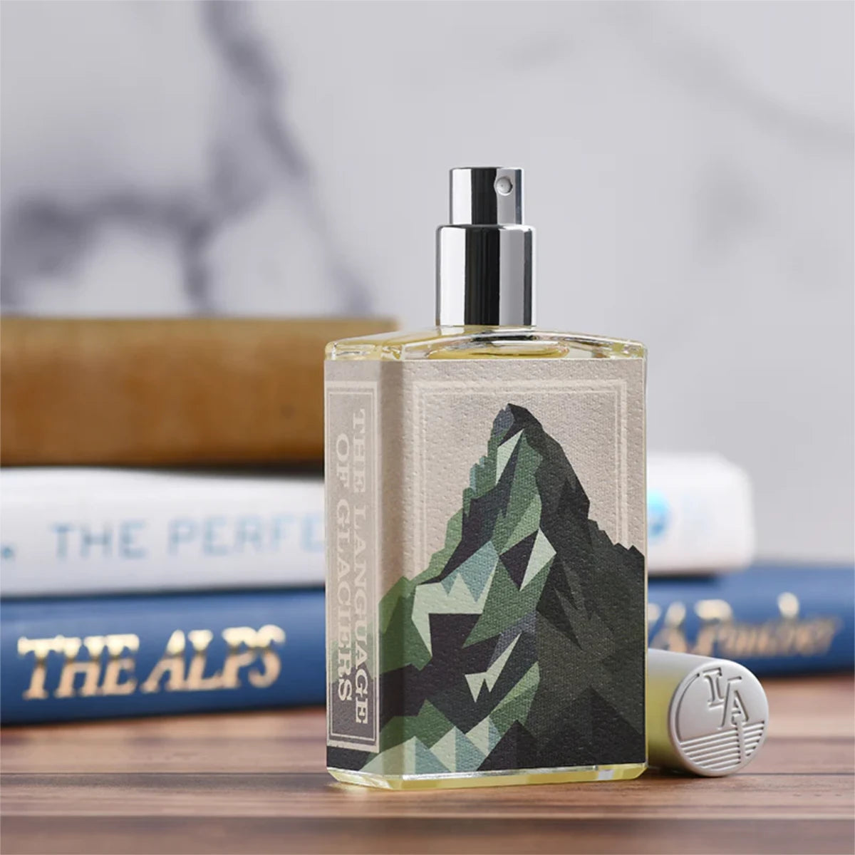 Imaginary Authors - The Language of Glaciers Parfumflakon mit geometrischem Bergmotiv vor unscharfem Bücherhintergrund