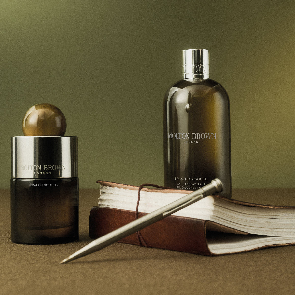 Molton Brown - Tobacco Absolute Parfumflakon und Duschgel daneben Lederbuch und silberner Stift auf grünem Untergrund