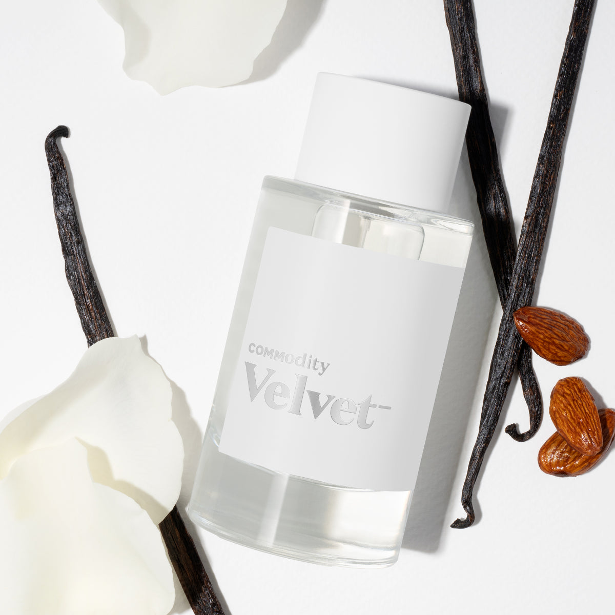 Commodity - Velvet- Duftflakon mit weißem Etikett, umgeben von Vanilleschoten und Blütenblättern auf hellem Hintergrund