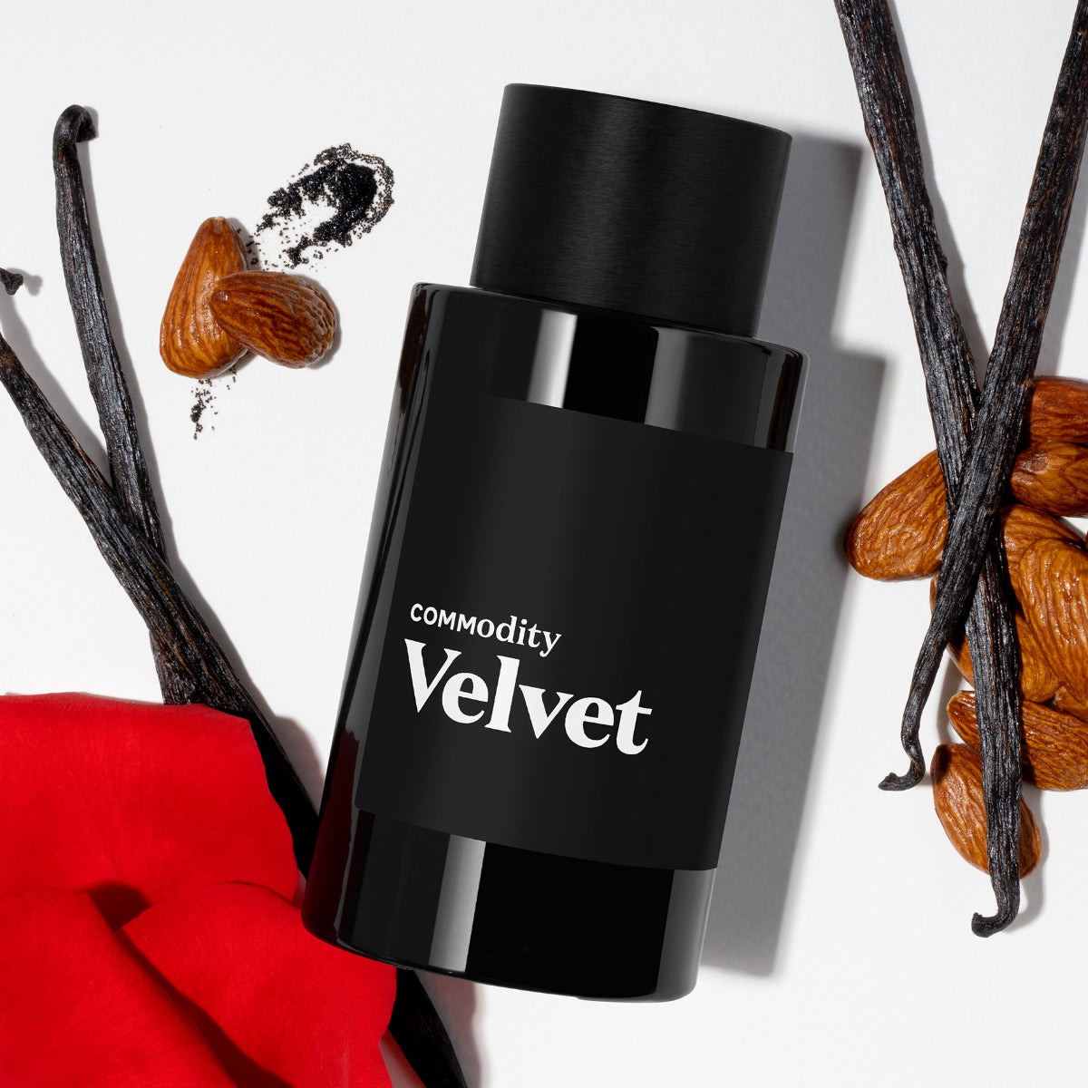 Commodity - Velvet Parfumflakon in schwarzem Design umgeben von Mandeln, Vanilleschoten und roter Rosenblüte auf hellem Untergrund