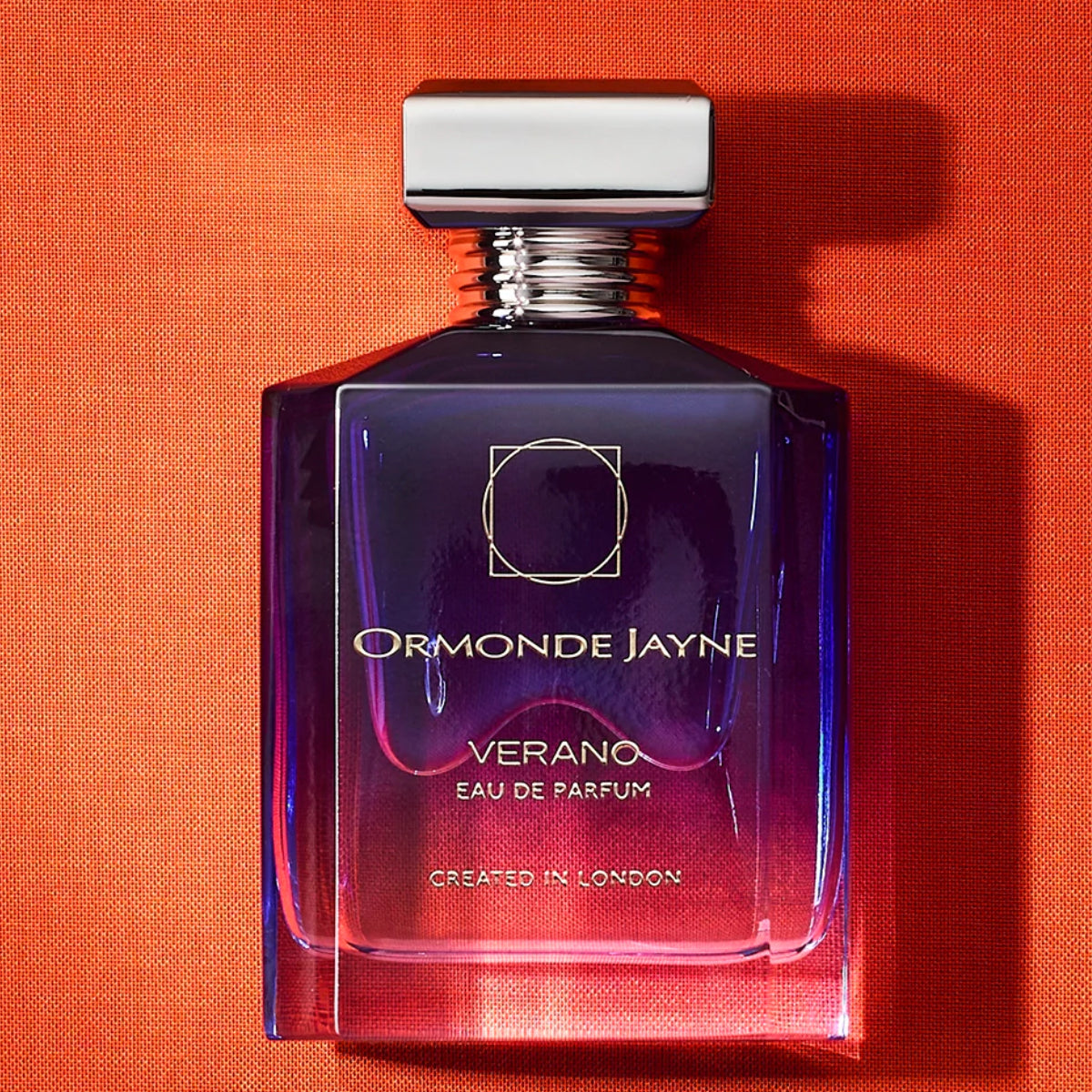 Ormonde Jayne - Verano Parfümflakon in tiefem Blau-Violett mit silberner Kappe vor orangem Hintergrund