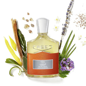 Creed - Viking Cologne Parfumflakon mit orangefarbenem Lederband und aromatischen Pflanzenarrangement auf weißem Untergrund