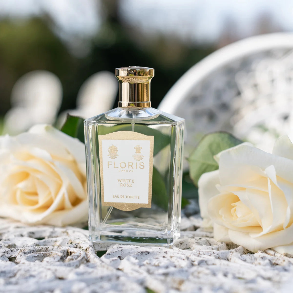 Floris - White Rose eleganter klarer Flakon mit goldfarbenem Verschluss umgeben von weißen Rosen auf Gartentisch