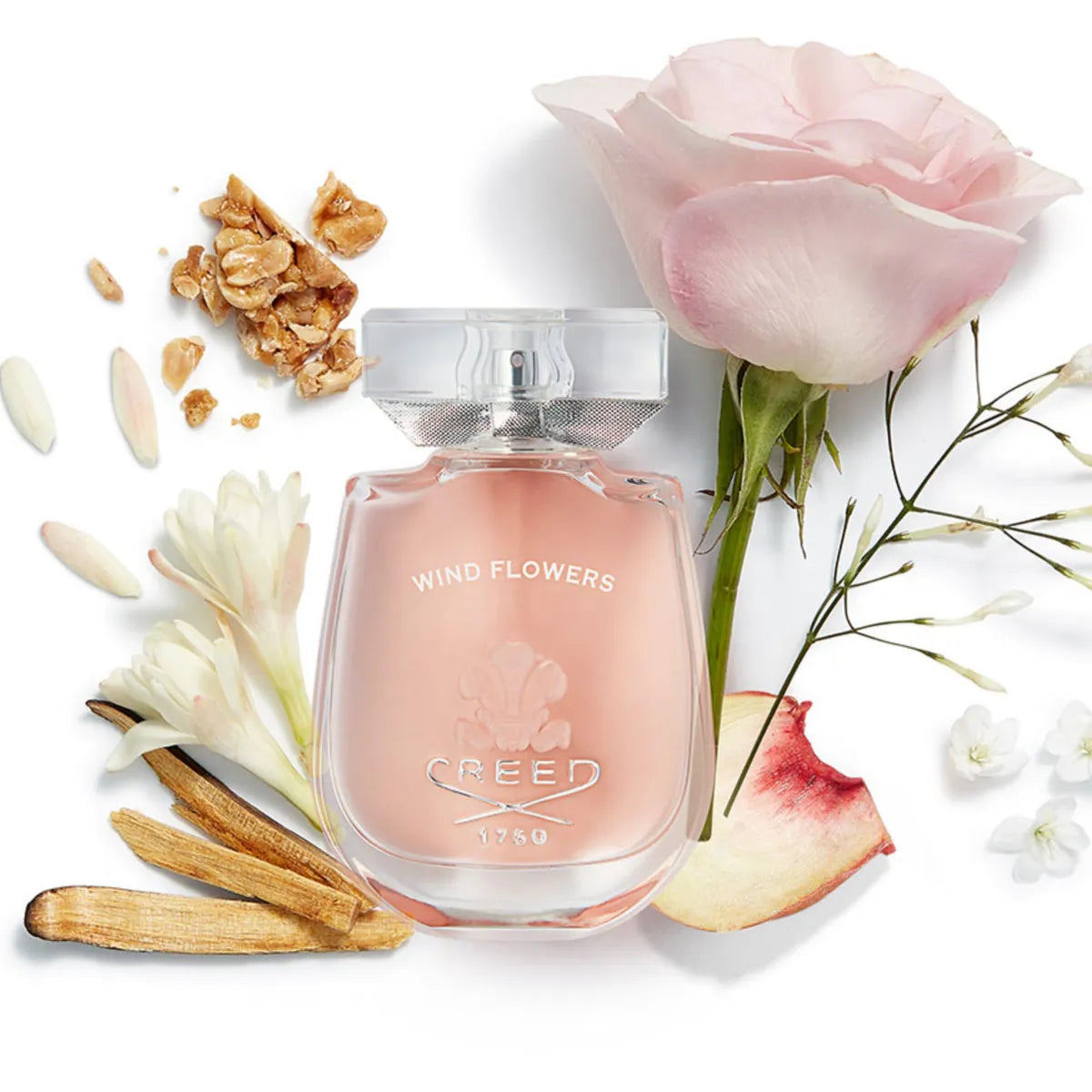 Creed - Wind Flowers Parfumflakon mit zart rosafarbenem Duft umgeben von Blüten und natürlichen Duftnoten vor weißem Hintergrund