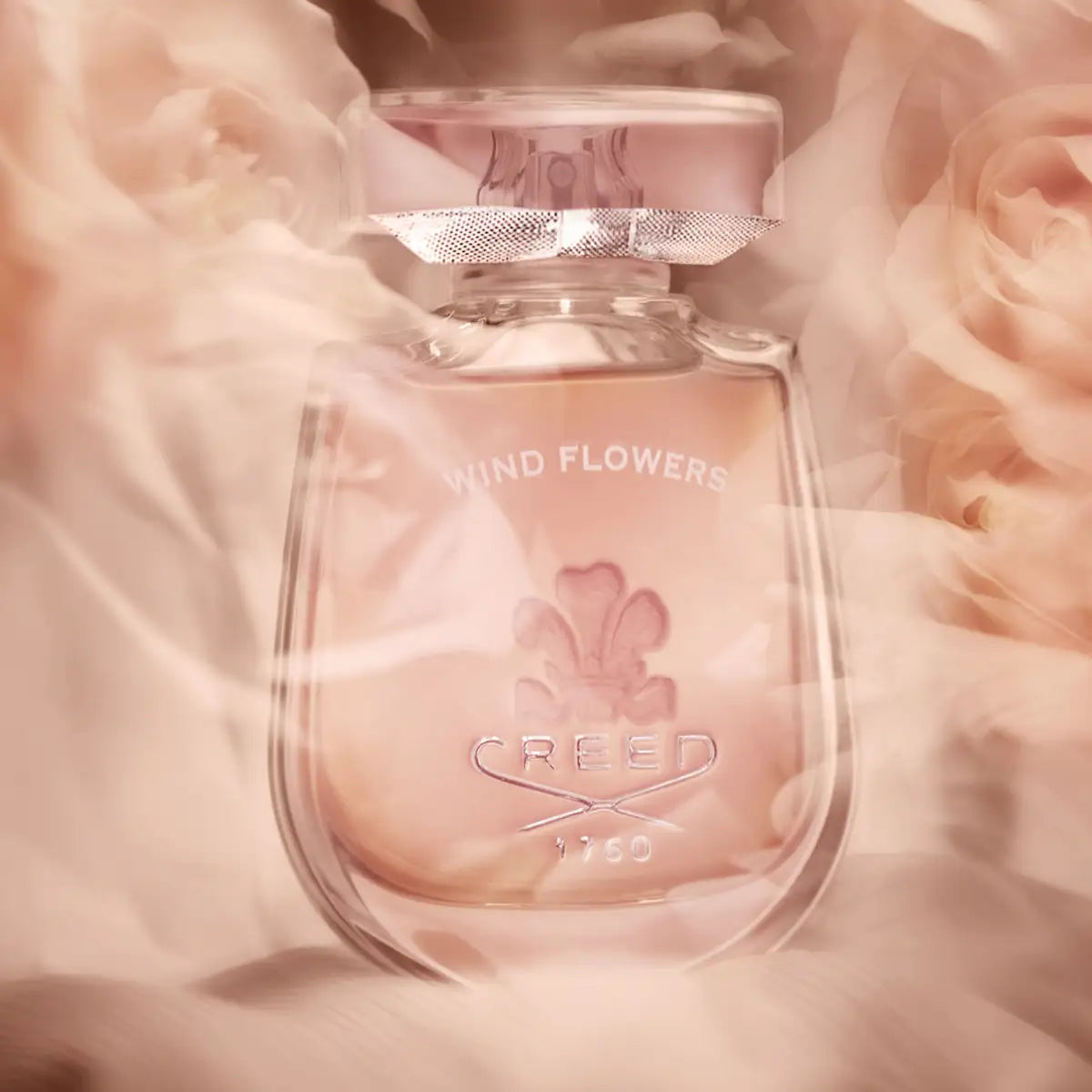Creed - Wind Flowers Parfumflakon mit zart rosa Flüssigkeit umgeben von weichen Blüten und Nebeleffekten