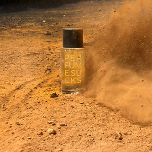PERFUME.SUCKS - Yellow 130C Flakon mit schwarzem Deckel und gelber Schrift in staubiger Wüstenlandschaft