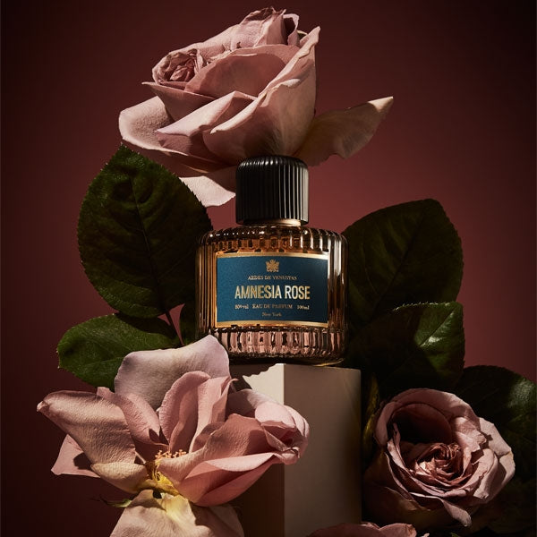 Aedes de Venustas - Amnesia Rose Parfumflakon mit geriffeltem Glas und blauem Etikett vor Rosen und dunklem Hintergrund