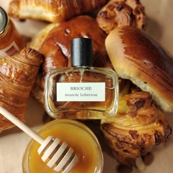 Anatole Lebreton - Brioche Duftflakon vor verschiedenen französischen Backwaren und Honigglas auf Holztisch