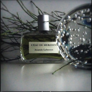 Anatole Lebreton - L’Eau de Merzhin Parfümflakon mit grünlicher Flüssigkeit und silbernem Verschluss vor dunklem Zweighintergrund