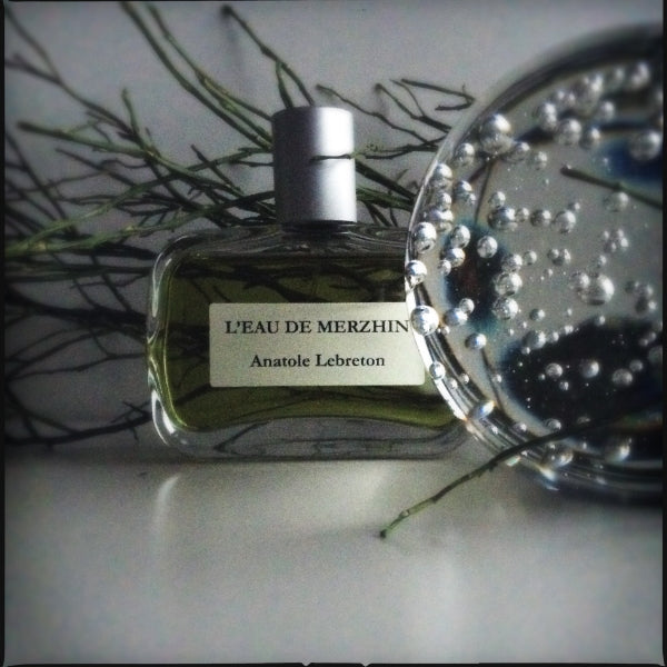 Anatole Lebreton - L’Eau de Merzhin Parfümflakon mit grünlicher Flüssigkeit und silbernem Verschluss vor dunklem Zweighintergrund