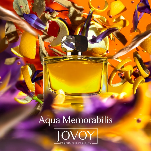 Jovoy - Aqua Memorabilis leuchtend goldgelber Flakon mit facettiertem Verschluss vor farbenfrohem, dynamischem Blüten- und Fruchtarrangement