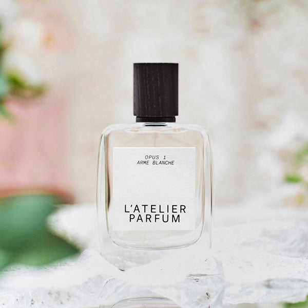 L'Atelier Parfum - Opus 1 - Arme Blanche klare Flasche mit schwarzem Holzverschluss vor sanft verschwommenem floralem Hintergrund