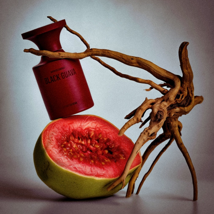 BORNTOSTANDOUT - Black Guava Flakon in dunklem Rot balanciert auf halbierter Guave mit geschwungenem Holzstück
