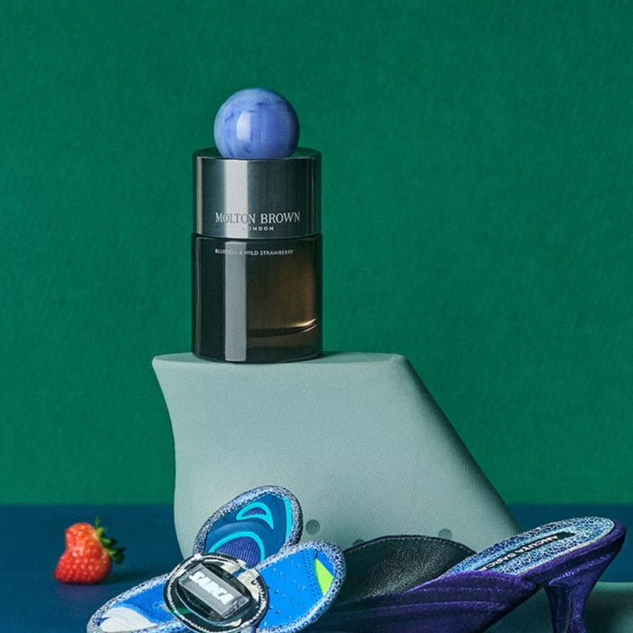 Molton Brown - Bluebell & Wild Strawberry zeigt dunklen Flakon mit blauem Kugelverschluss vor grünem Hintergrund