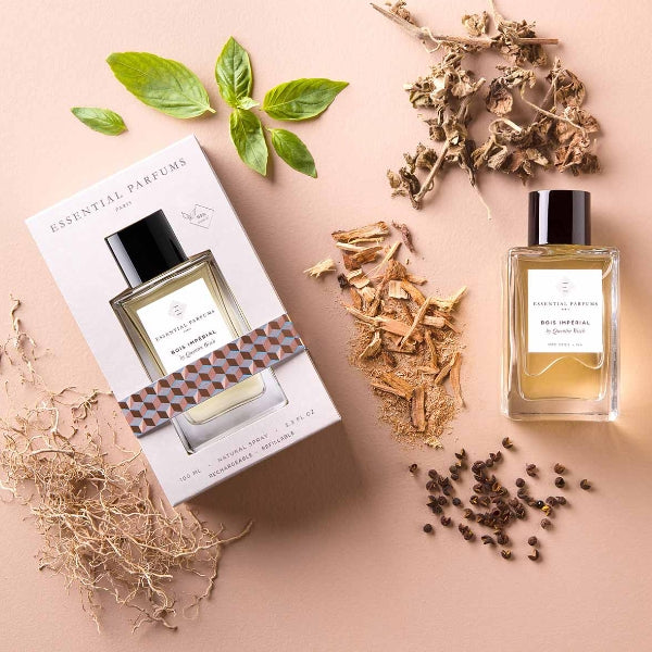 Essential Parfums - Bois Impérial Flakon mit Verpackung umgeben von Kräutern und Holzstücken auf hellem Untergrund