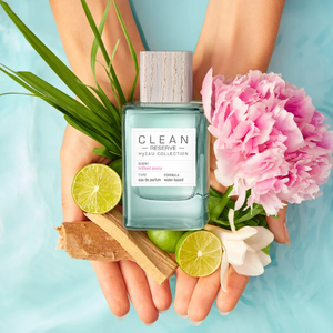 Clean Perfume - H2Eau Brilliant Peony Flakon mit Holzdeckel, Pfingstrose, Limetten und Grünpflanze in Händen vor blauem Hintergrund