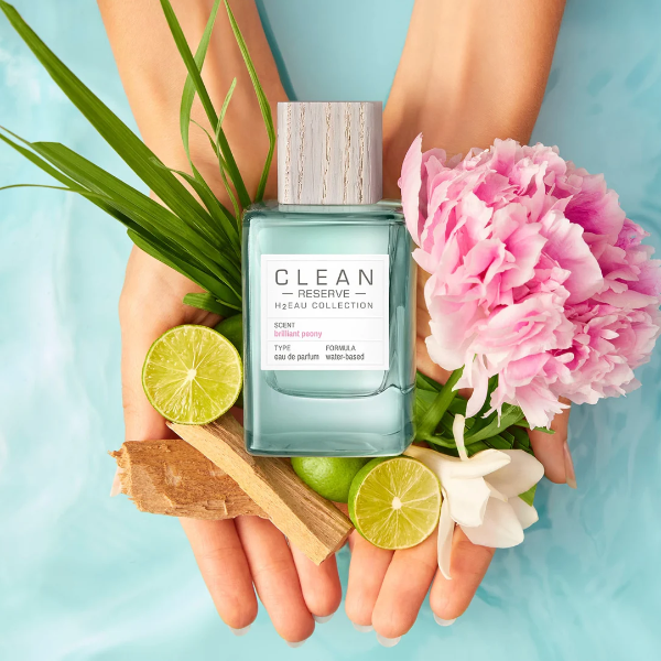 Clean Perfume - H2Eau Brilliant Peony Flakon mit Holzdeckel, Pfingstrose, Limetten und Grünpflanze in Händen vor blauem Hintergrund