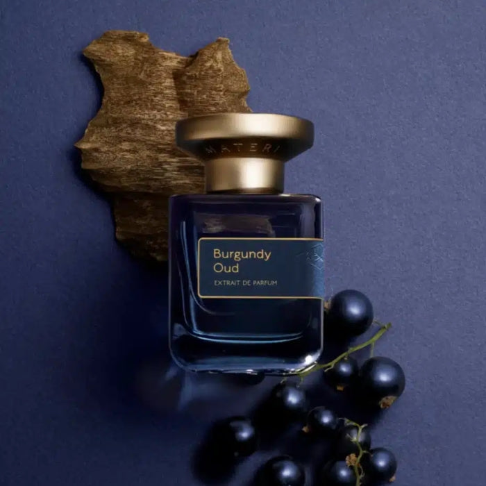 Atelier Materi - Burgundy Oud Parfumflakon in tiefblauem Glas mit goldenem Verschluss vor dunklem Holz und Beeren