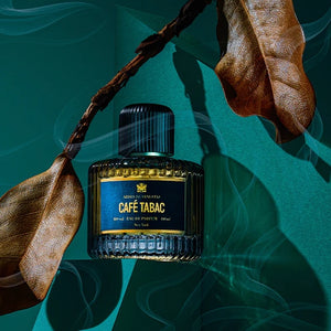Aedes de Venustas - Café Tabac Parfumflakon mit dunkelblauem Etikett zwischen getrockneten Blättern und Rauch auf grünem Hintergrund