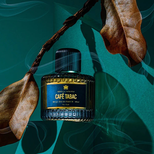Aedes de Venustas - Café Tabac Parfumflakon mit dunkelblauem Etikett zwischen getrockneten Blättern und Rauch auf grünem Hintergrund