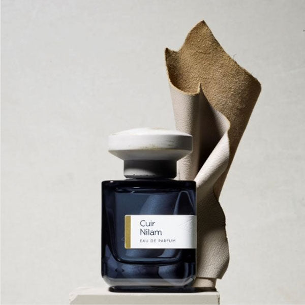 Atelier Materi - Cuir Nilam Parfumflakon in dunklem Glas mit hellem, rundem Verschluss vor Lederskulptur