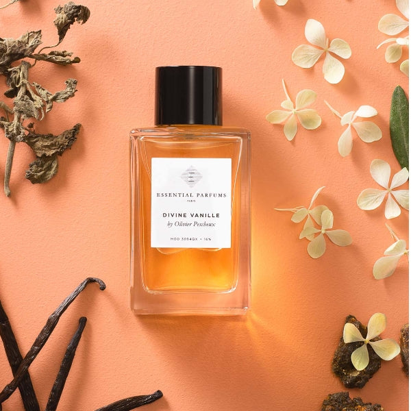 Essential Parfums - Divine Vanille Parfümflakon mit schwarzem Verschluss auf apricotfarbenem Hintergrund mit Vanillestangen und Blüten