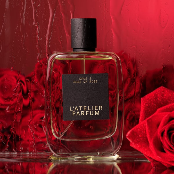 L'Atelier Parfum - Opus 2 - Dose of Rose klarer Flakon mit schwarzem Etikett vor rotem Hintergrund und Rosenblüten