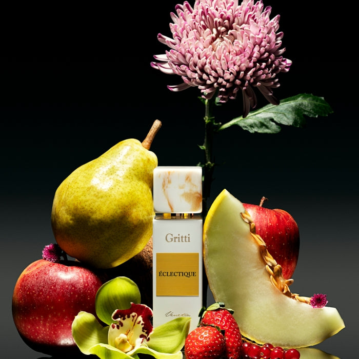 Gritti - White Collection - Eclectique zeigt eleganten weißen Flakon mit goldfarbenem Etikett umgeben von Früchten und Blüten vor dunklem Hintergrund