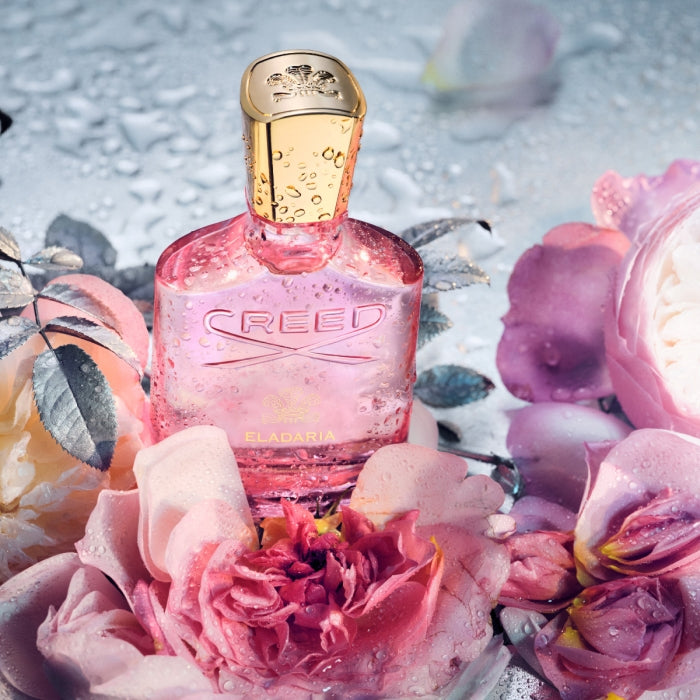 Creed - Eladaria Flakon in rosa Glas mit goldfarbenem Verschluss umgeben von taufrischen Rosenblüten und Blättern