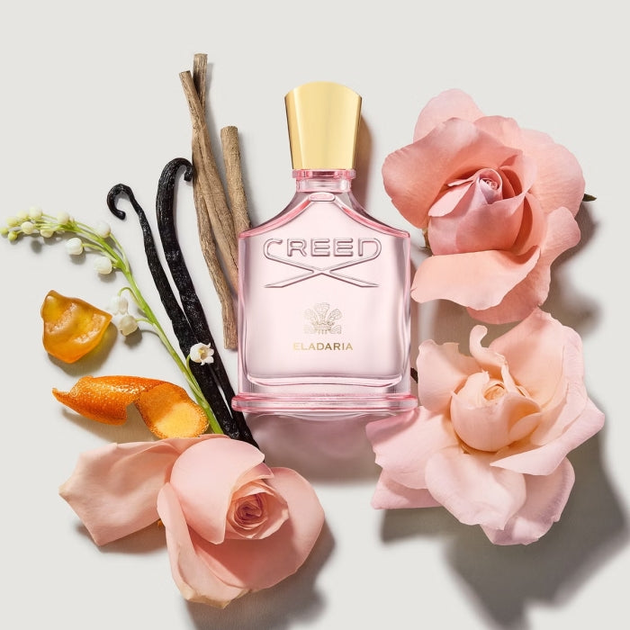 Creed - Eladaria Parfumflakon mit zartrosa Glas und goldfarbenem Verschluss umgeben von Rosen und Vanilleschoten