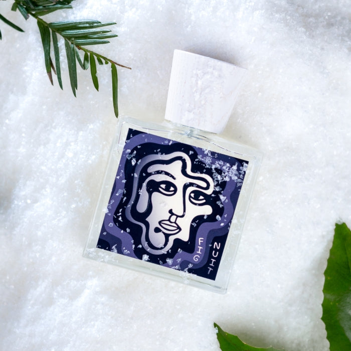 Carl Kling Parfums - Fig Nuit Flakon mit abstrakter Gesichtsgrafik auf dunklem Hintergrund liegt auf Schnee vor grünem Blatt