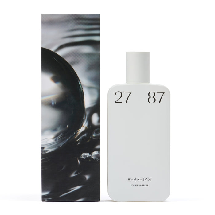 27 87 - #hashtag Parfumflakon in schlichtem Weiß mit klarer Typografie neben künstlerischer Verpackung vor weißem Hintergrund