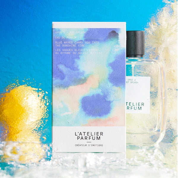 L'Atelier Parfum - Opus 3 - Hot Splash Verpackung mit blauem Farbverlauf neben klarem Flakon vor blau-gelbem Hintergrund