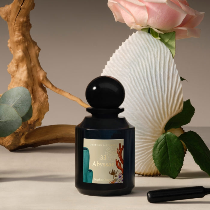 L'Artisan Parfumeur - La Botanique - Abyssae Parfumflakon mit schwarzem Glas und dekorativem Etikett vor natürlichem Arrangement