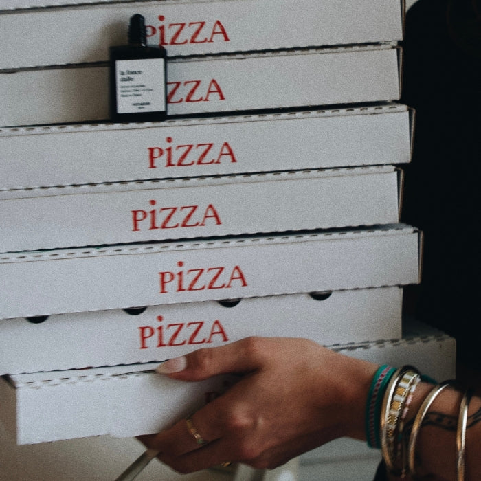 Versatile - La Foncedalle Roll-on Parfumflakon auf gestapelten Pizzakartons mit Hand und Armbändern in Alltagsszene