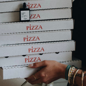 Versatile - La Foncedalle Roll-on Parfumflakon auf gestapelten Pizzakartons mit Hand und Armbändern in Alltagsszene