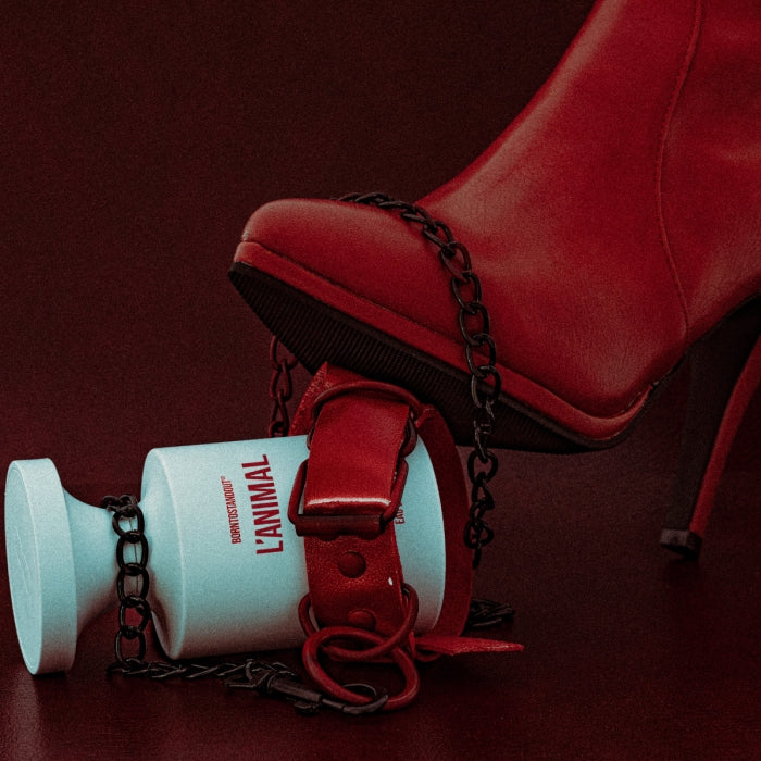 BORNTOSTANDOUT - L'Animal weißer Parfumflakon mit roter Schrift, kombiniert mit rotem Schuh und schwarzer Kette vor dunkelrotem Hintergrund